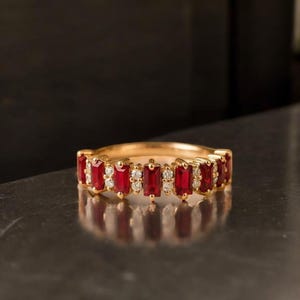 May include: A gold ring featuring alternating rectangular red gemstones and small white gemstones. The ring has a polished gold band and is set against a dark background, highlighting the vibrant colors of the gemstones.