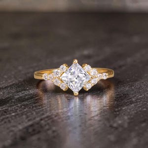 Peut inclure: Une bague en or ornée d'une pierre précieuse carrée et transparente au centre, entourée de petites pierres rondes. Le corps de la bague est en or uni, et le sertissage présente des grappes de petites pierres de chaque côté du joyau central.