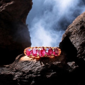 Puede incluir: Un anillo de oro con cinco piedras preciosas rojas de corte ovalado. El anillo se muestra sobre una roca marrón oscura texturizada, con un fondo brumoso azul claro. Las piedras están espaciadas uniformemente y engastadas en la banda de oro.