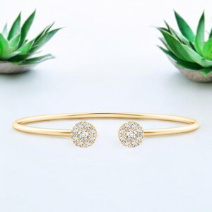 Pode incluir: Uma pulseira de punho aberta dourada. Apresenta dois grupos circulares de pequenas pedras preciosas transparentes. A pulseira tem um design simples e elegante. As pedras preciosas estão dispostas em um padrão floral. O fundo é branco.