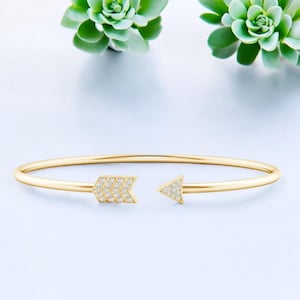 Może przedstawiać: Złota bransoletka w kształcie strzały o delikatnym wzorze. Groty strzał zdobione są małymi, błyszczącymi kamieniami. Bransoletka umieszczona jest na miękkim, jasnym tle, z zielonymi roślinami w tle.