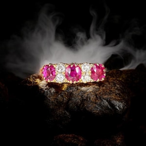 May include: A gold ring set with five oval-cut, vivid pink gemstones, interspersed with smaller, round, clear gemstones. The ring is displayed on a dark, textured surface, against a smoky, dark backdrop.