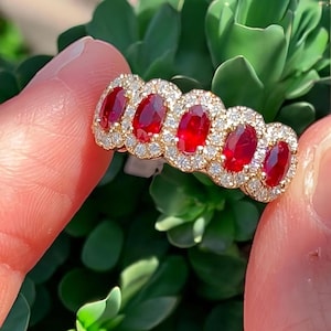 May include: A gold ring featuring five oval-cut, deep red gemstones, each surrounded by a halo of smaller, clear gemstones. The ring is held against a backdrop of green foliage, highlighting the ring's sparkle.