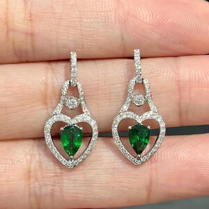 May include: A pair of silver earrings with a heart-shaped design. Each earring features a pear-shaped, vibrant green gemstone at the center of the heart, surrounded by small, sparkling stones. The earrings are held up by a small, round stone.