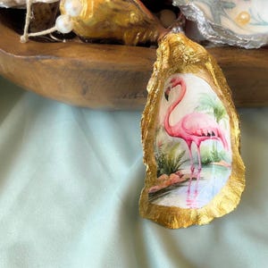 May include: An oyster shell with a gold leaf border features a pink flamingo illustration. The flamingo stands in a tropical scene with green foliage and water. The shell is set against a light green background.