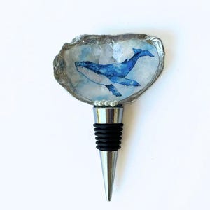 May include: A decorative bottle stopper featuring a blue whale painted inside a shell. The shell has a silver-coloured edge and is mounted on a silver and black stopper with a pearl accent. The whale is painted in shades of blue.