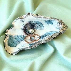 May include: An oyster shell jewelry dish with a pearl and two rings. The dish has a white interior with blue and gray painted details. The shell's edge is silver. The rings are gold-colored.