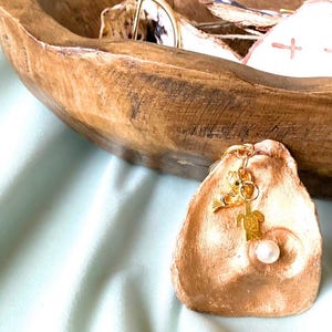 May include: A gold-coloured charm with a turtle and fish design, paired with a white pearl, hangs from a small, tan-coloured, seashell-shaped dish. The dish sits on a light blue surface, with a wooden bowl in the background.