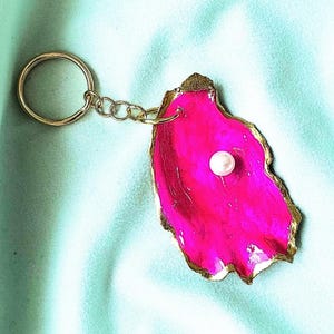 May include: A vibrant pink oyster shell keyring with a gold-coloured edge and a small white pearl. The keyring has a gold-coloured ring and chain. The background is a light blue fabric.