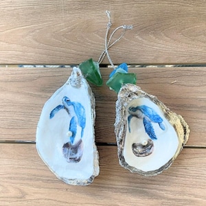 May include: Two oyster shell ornaments with a blue sea turtle design. Each shell has a pearl and is topped with a piece of sea glass. Silver string is attached for hanging. The shells are on a wooden surface.