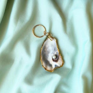 May include: A unique keychain featuring a natural oyster shell with a gold-colored rim and a small pearl. The shell is attached to a gold-colored key ring. The background is a light green fabric.