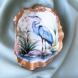 May include: An oyster shell dish with a gold-painted edge. The interior features a painted scene of a blue heron standing in water, surrounded by plants and rocks. The heron has blue and white feathers and red legs.