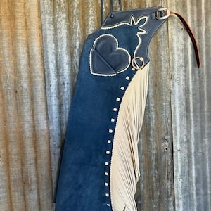 May include: Navy blue suede chaps with a heart-shaped leather patch and cream-coloured fringe. The chaps feature a decorative design along the top edge and are accented with white stitching and a leather belt.