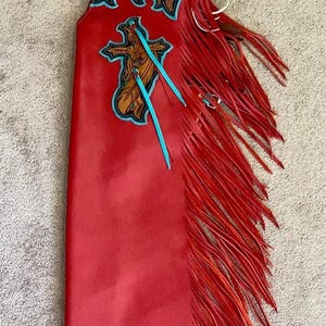 Könnte beinhalten: Rote Lederchaps mit aufwendigen Verzierungen und Fransen. Verfügt über ein Kreuzdesign mit türkisfarbenen Akzenten und einen braunen Ledergürtel. Ein Kleidungsstück im Western-Stil.
