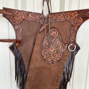 May include: Brown leather chaps with detailed floral tooling. The chaps have a suede-like texture, intricate floral designs, and long fringe. Metal ring and leather straps are visible. The chaps are designed for riding.