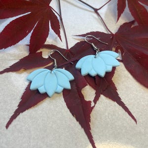 May include: A pair of light blue lotus flower earrings with silver-colored hooks. The earrings are set against a backdrop of deep red maple leaves, creating a striking contrast of colors and textures. The earrings are a delicate, floral-themed accessory.