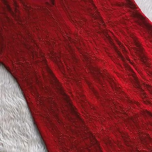May include: A vibrant red tunic with intricate embroidered detailing. The long-sleeved garment features a round neckline and a delicate floral pattern throughout. The tunic is displayed on a wooden hanger.