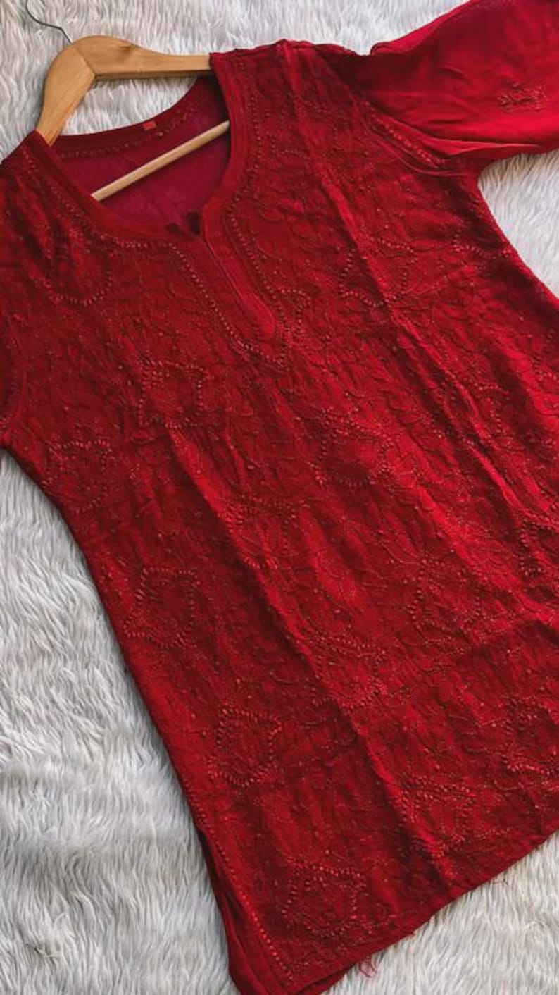May include: A vibrant red tunic with intricate embroidered detailing. The long-sleeved garment features a round neckline and a delicate floral pattern throughout. The tunic is displayed on a wooden hanger.