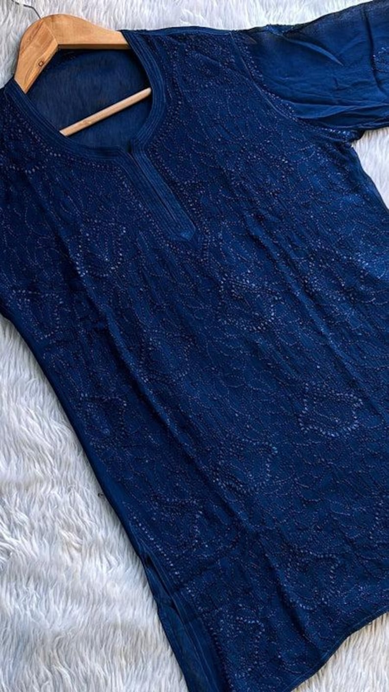 May include: A navy blue tunic top with intricate embroidered detailing. The top features a round neckline with a small slit and short sleeves. The fabric appears lightweight and is displayed on a wooden hanger.
