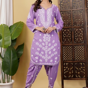 Puede incluir: Un conjunto de salwar kameez lavanda con intrincados bordados blancos. La túnica de manga larga y los pantalones a juego presentan patrones florales y geométricos. El atuendo se usa con sandalias blancas. Una pantalla de madera y una planta en maceta están en el fondo.