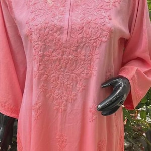 Puede incluir: Una t&uacute;nica de color coral claro con bordado floral. La parte superior de manga larga presenta costuras detalladas alrededor del cuello y en la parte delantera. La tela parece suave y ligera, adecuada para climas c&aacute;lidos.