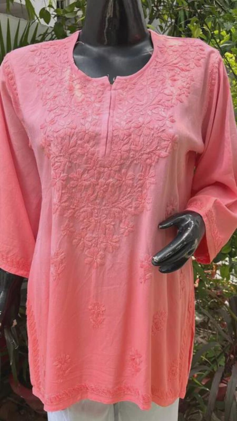 Puede incluir: Una t&uacute;nica de color coral claro con bordado floral. La parte superior de manga larga presenta costuras detalladas alrededor del cuello y en la parte delantera. La tela parece suave y ligera, adecuada para climas c&aacute;lidos.