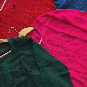 May include: Four embroidered tunics in shades of red, blue, pink, and green, displayed on wooden hangers. The fabric features detailed floral patterns. The tunics are made of a lightweight material.