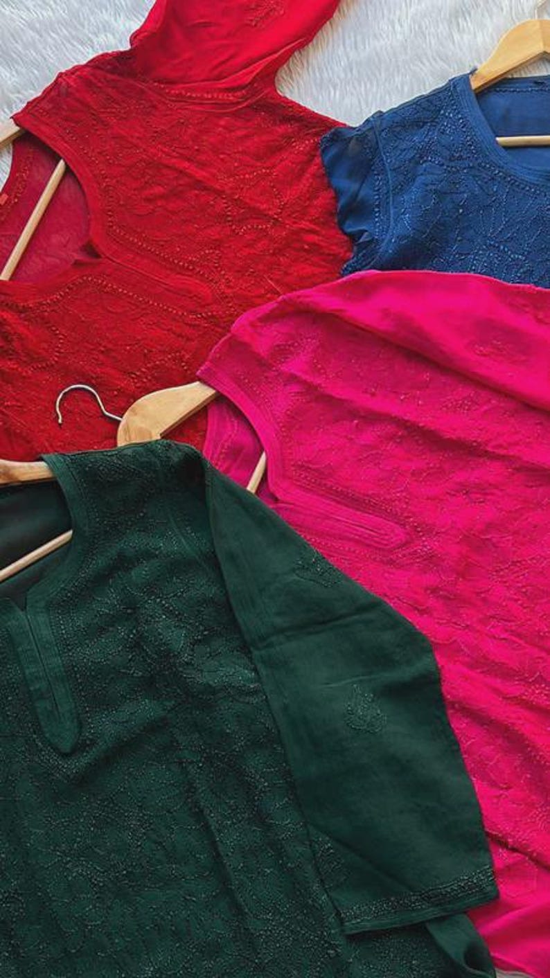 May include: Four embroidered tunics in shades of red, blue, pink, and green, displayed on wooden hangers. The fabric features detailed floral patterns. The tunics are made of a lightweight material.