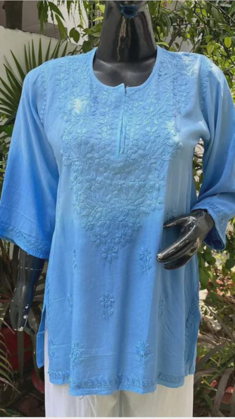 Puede incluir: Una t&uacute;nica azul claro con intrincados bordados florales. La t&uacute;nica presenta un cuello redondo, mangas tres cuartos y aberturas laterales. El bordado se concentra en el pecho y el dobladillo inferior, creando un dise&ntilde;o delicado y texturizado.