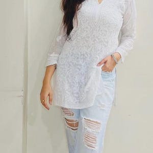 Puede incluir: Túnica blanca bordada con cuello en V y mangas tres cuartos. La túnica se combina con vaqueros rotos de color azul claro. El conjunto se completa con zapatos rojos. El estilo general es casual y chic.