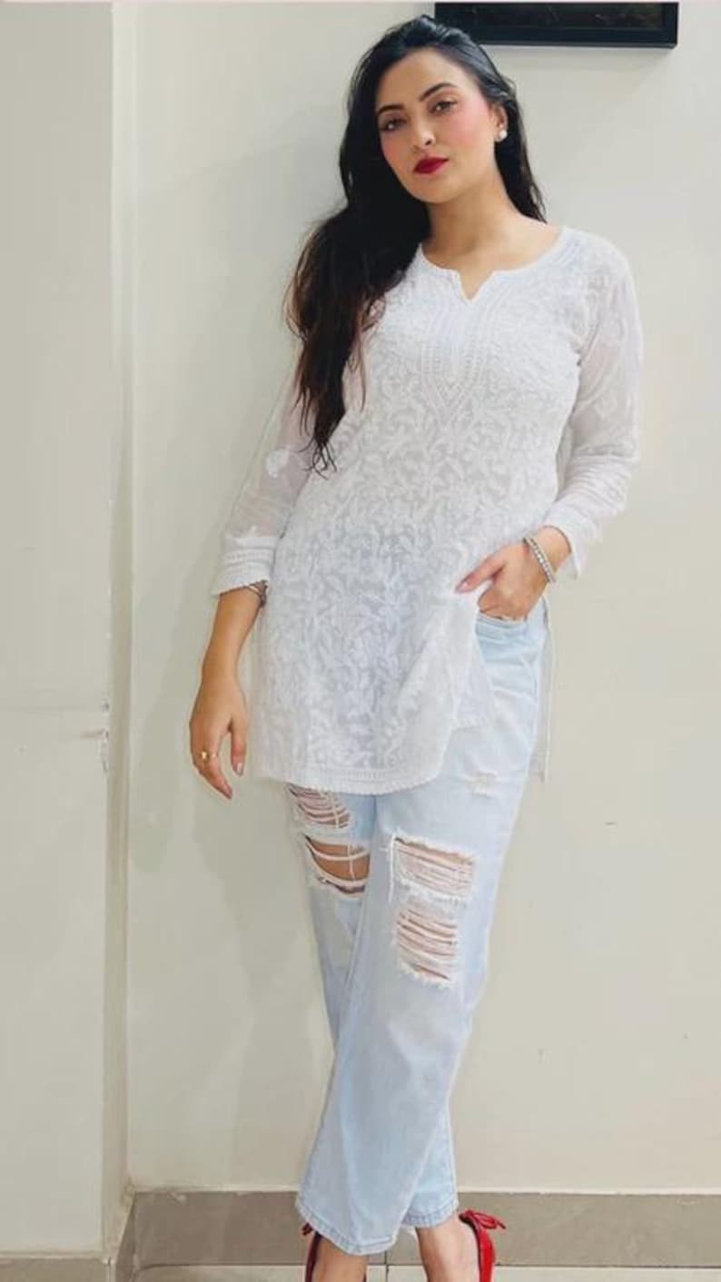 May include: White embroidered tunic with a V-neck and three-quarter sleeves. The tunic is paired with light blue ripped jeans. The outfit is completed with red shoes. The overall style is casual and chic.
