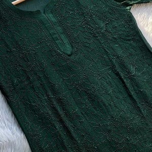 May include: Dark green tunic top with intricate embroidered detailing. The top features a round neckline with a small slit and long sleeves. The fabric appears to be lightweight and flowing, with a subtle sheen. The garment is displayed on a wooden hanger.