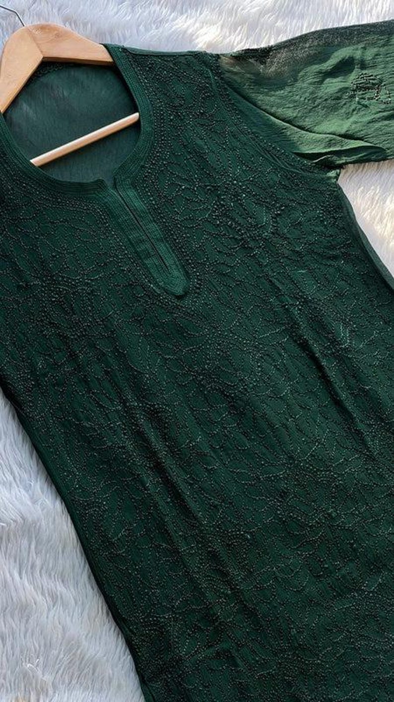 May include: Dark green tunic top with intricate embroidered detailing. The top features a round neckline with a small slit and long sleeves. The fabric appears to be lightweight and flowing, with a subtle sheen. The garment is displayed on a wooden hanger.