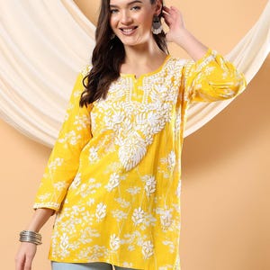 May include: A yellow tunic with white floral embroidery and a round neckline. The three-quarter sleeve top features a floral pattern and is paired with light blue jeans. The tunic is made of a lightweight fabric.