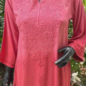 Puede incluir: T&uacute;nica de color coral con intrincados bordados florales. Las mangas tres cuartos y el escote tambi&eacute;n est&aacute;n bordados. La t&uacute;nica se exhibe en un maniqu&iacute; negro. El fondo incluye follaje verde.