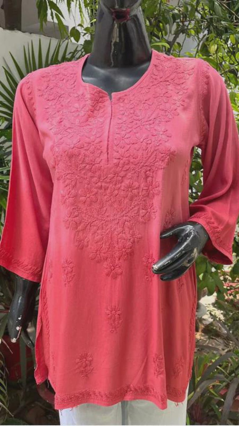 Puede incluir: T&uacute;nica de color coral con intrincados bordados florales. Las mangas tres cuartos y el escote tambi&eacute;n est&aacute;n bordados. La t&uacute;nica se exhibe en un maniqu&iacute; negro. El fondo incluye follaje verde.