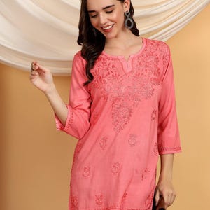 May include: A coral-colored tunic with three-quarter sleeves and intricate floral embroidery around the neckline and bodice. The tunic has a relaxed fit and a subtle V-neck. The model is wearing dark blue pants and holding a brown wallet.