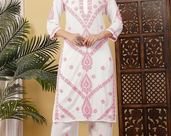 May include: A white long-sleeved tunic and wide-leg trousers set featuring intricate pink floral embroidery throughout. The tunic has a round neckline with a small vertical slit. The logo "ADA" is visible in the top left corner of the image.