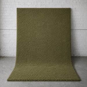 May include: Olive green rectangular rug with a textured surface. The rug is displayed against a white brick wall and a gray concrete floor. The rug is folded over at the bottom.