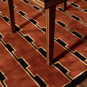 May include: A wooden table with a burl wood top and striped legs stands on a rug featuring a repeating geometric pattern of terracotta red, black, and gold lines. The table legs are constructed from alternating light and dark wood strips.