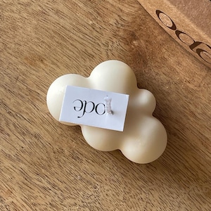May include: A cream-coloured cloud-shaped candle with a white label reading "ode" in black script. The candle is on a wooden surface, with a partially visible cardboard box in the background.