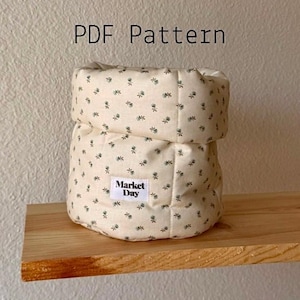 Może przedstawiać: Beżowy pojemnik z tkaniny z kwiatowym wzorem i napisem "Market Day" na małej etykiecie. Pojemnik stoi na drewnianej półce. Tekst "PDF Pattern" znajduje się powyżej.