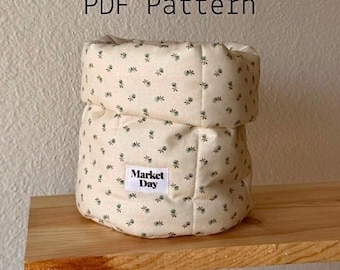 May include: A beige fabric container with a floral pattern and the text "Market Day" on a small label. The container is sitting on a wooden shelf. The text "PDF Pattern" is above the container.