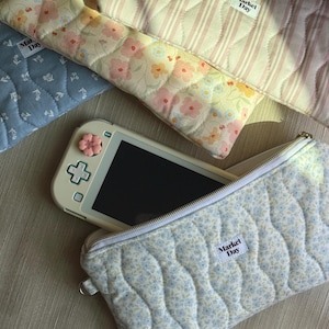 Peut inclure: Plusieurs pochettes en tissu matelassé à motifs floraux et rayés. L'une d'elles est ouverte, révélant une console de jeu blanche avec un bouton en forme de fleur rose. Les pochettes ont un motif matelassé ondulé et une étiquette "Market Day".