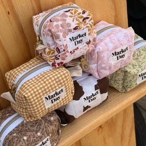 May include: A stack of small, square fabric pouches with zippered closures. The pouches feature various patterns, including floral, gingham, and a cowhide print. Each pouch has a white label that reads "Market Day".