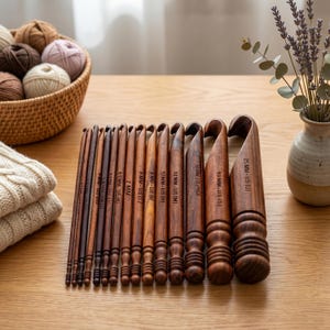 Può includere: Una collezione di uncinetti in legno di varie dimensioni, disposti su una superficie di legno. Gli uncinetti variano in dimensioni da 5 mm a 25 mm, con le dimensioni stampate su ogni uncinetto. Un cesto di filato e un vaso di fiori sono sullo sfondo.