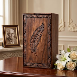 May include: A dark brown wooden urn featuring a carved feather design. The rectangular urn has a leaf border. A framed photograph and a bouquet of white flowers are placed nearby. The urn is likely for ashes.