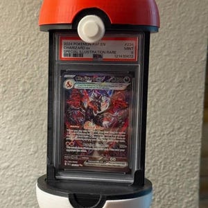 May include: A red and white 3D-printed Poké Ball display case holding a graded Pokémon card. The card features a Charizard ex illustration, with text indicating it's a Special Illustration Rare from 2024.