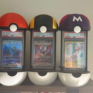 Pode incluir: Tr&ecirc;s expositores de cartas Pok&eacute;mon. Cada suporte apresenta um topo de cor diferente: vermelho, preto e amarelo, e roxo com um "M" branco. Os suportes t&ecirc;m uma base branca e cont&ecirc;m cartas Pok&eacute;mon classificadas.