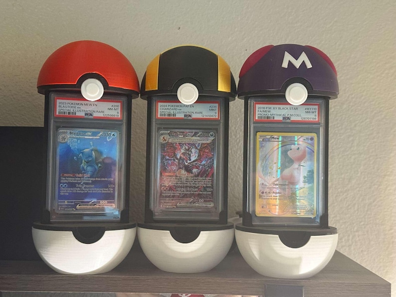 Pode incluir: Tr&ecirc;s expositores de cartas Pok&eacute;mon. Cada suporte apresenta um topo de cor diferente: vermelho, preto e amarelo, e roxo com um "M" branco. Os suportes t&ecirc;m uma base branca e cont&ecirc;m cartas Pok&eacute;mon classificadas.
